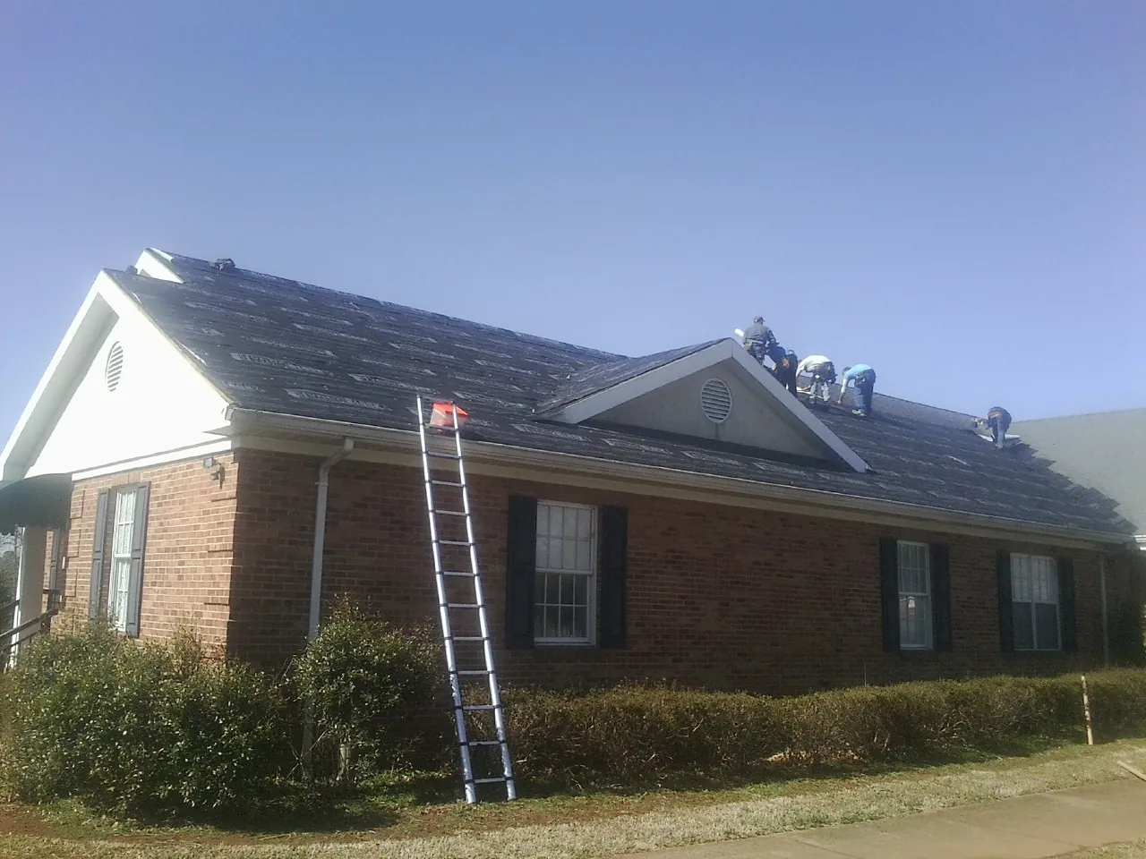 Crew tearing shingles off a brick ranch roof with a ladder set — Emergency Roof Repair work by our crew in Mont Belvieu
