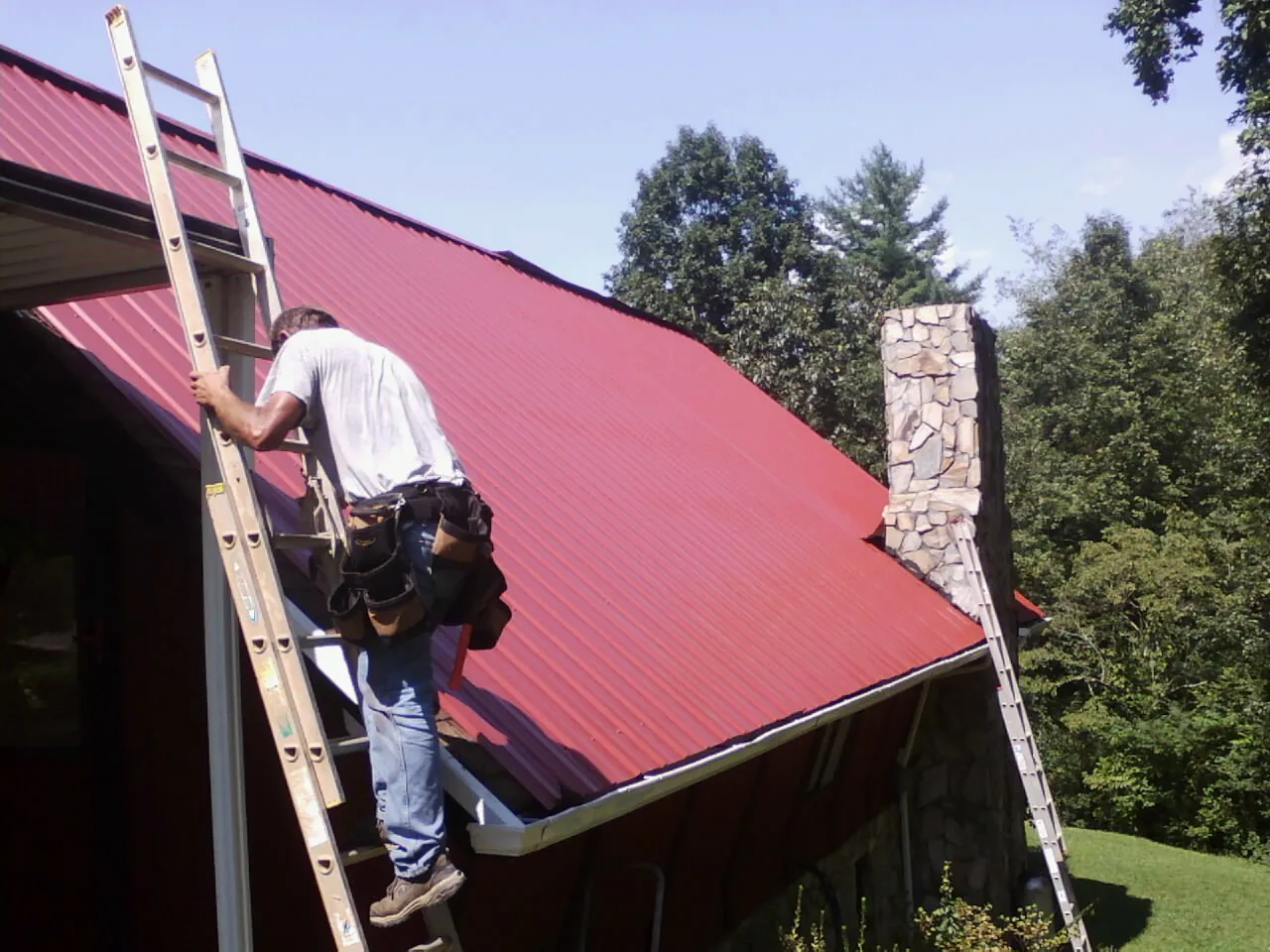 Roofer on a ladder beside a red metal roof and stone chimney — Soffit & Fascia Repair by Roofing Brothers in Mont Belvieu